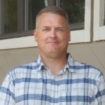 Jason with a blue and white checked shirt stands outside the Stayton office.