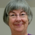 a head shot of Stephanie, smiling with lips shut, frameless glasses, sporting a salt and pepper bob hair cut and wearing a purple shirt.