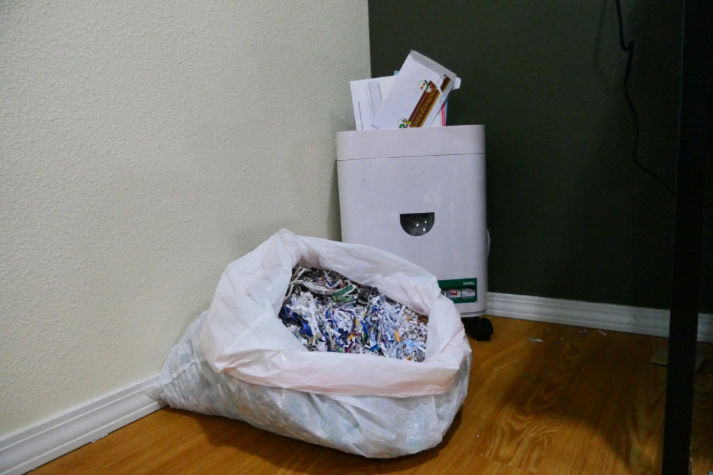 A large plastic garbage bag filled with shredded paper rests in front of a paper shredder with junk mail popping out the top. 