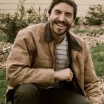 Kurt in a Carhartt jacket crouching down with his hand on a piece of wood.