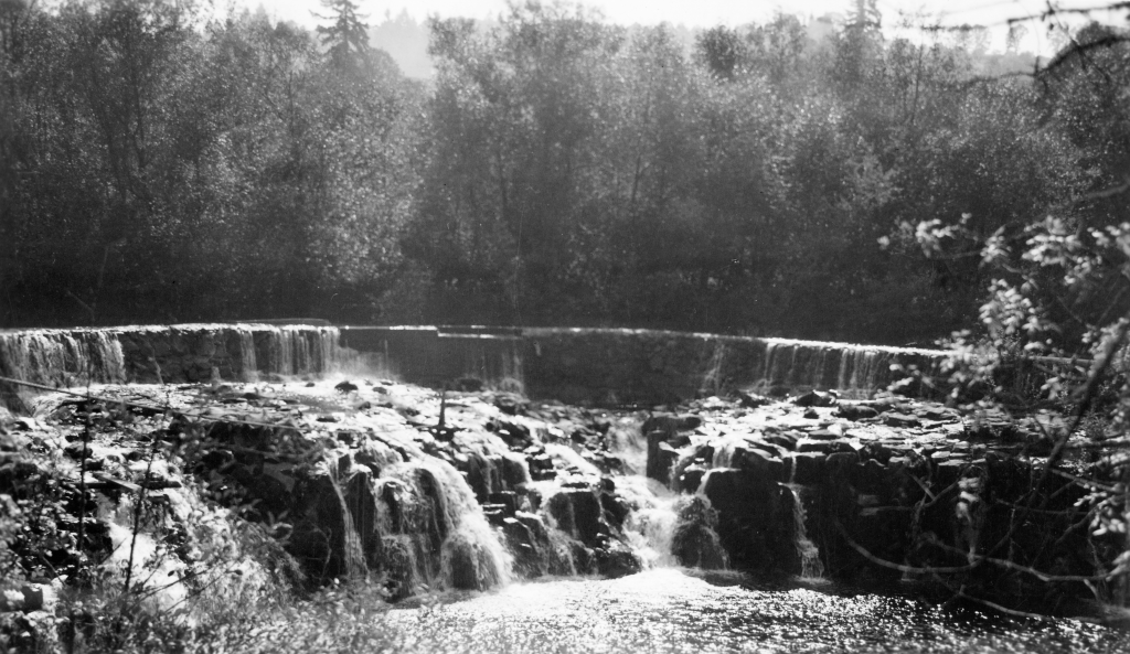 Historical black and white photo of Scotts Mills Dam showing water cascading over the top of the dam and then heading down over the waterfall.