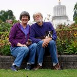a photo of Stephanie and Ray and their little black and white dog sitting on a garden bed edge outside the capital building in Salem, OR.