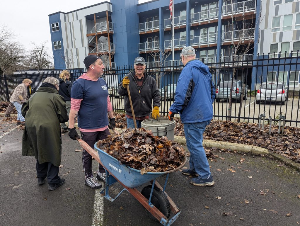 Man with wheelbarrow full of leaves and others around him tending to parking lot strip planting project.